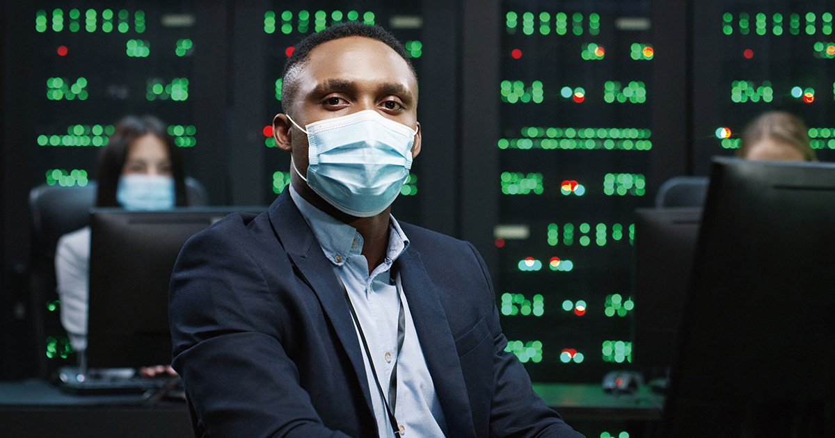 Close up of African American IT worker in mask working on desktop computers in data center control room and looking at camera Male security engineer working on computer. Female analytics on background