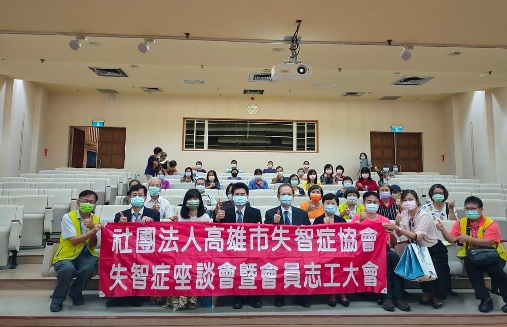 可能是 3 個人、大家站著和顯示的文字是「社團法人高雄市失智症協會 失智症座談會暨會員志工大會」的圖像
