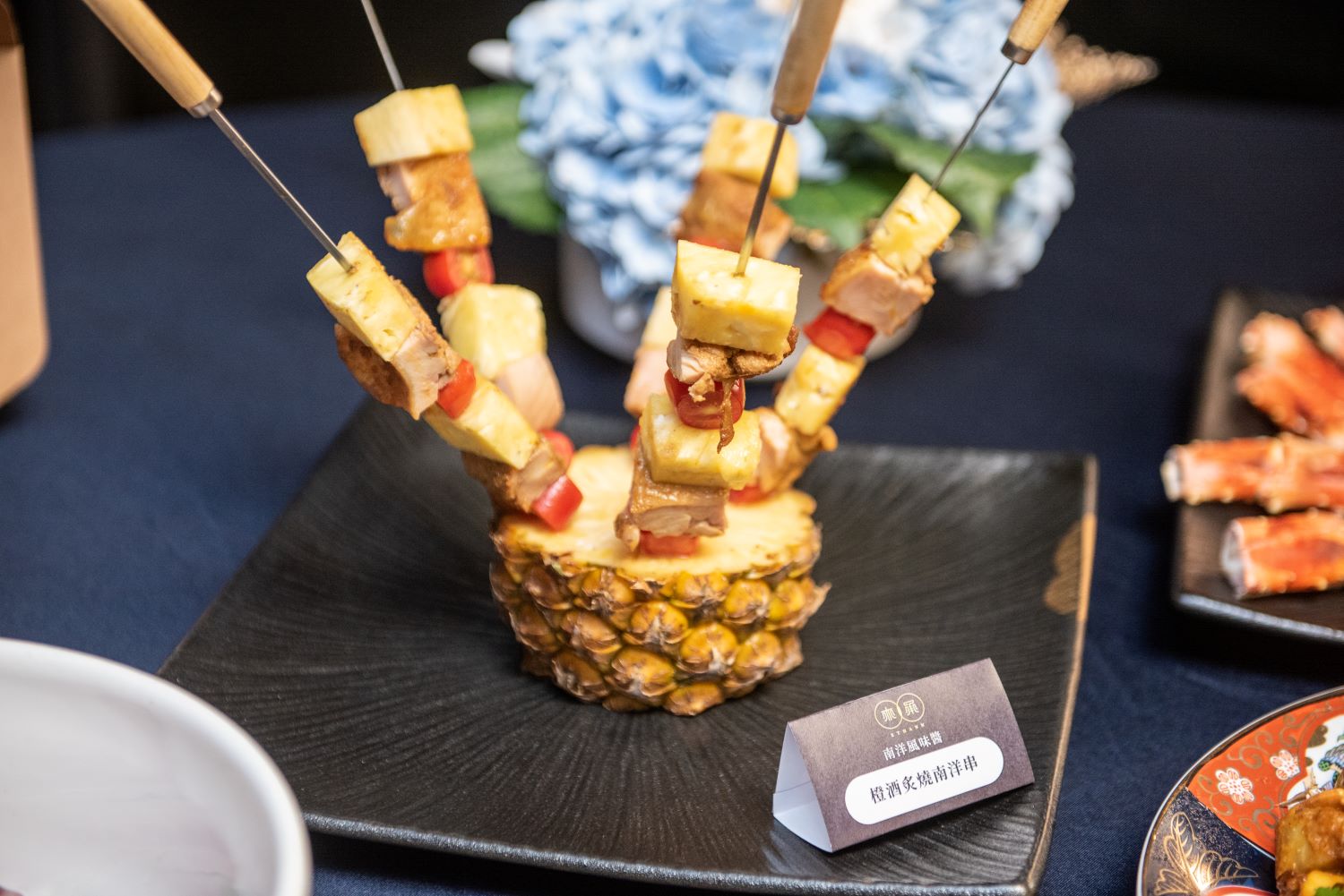 「橙酒炙燒南洋串」使用南洋風味醬醃漬去骨雞腿肉、鳳梨、番茄並燒烤,最後為了增加焦香味,上桌時還會用橙酒與高粱酒淋上烤肉串炙燒增添香氣。(圖/業者提供)