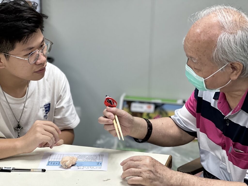 學生(左)向標準病人(右)說明為他量身訂做的筷子輔助器,作品獲113年台灣職能治療學會輔具競賽銀牌。(圖/中山醫大提供)