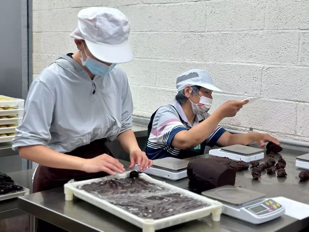 單親跨代照顧者囿囿(右)在烘焙坊協助餡料秤重,邊學邊做,重拾生活節奏。(芥菜種會提供)