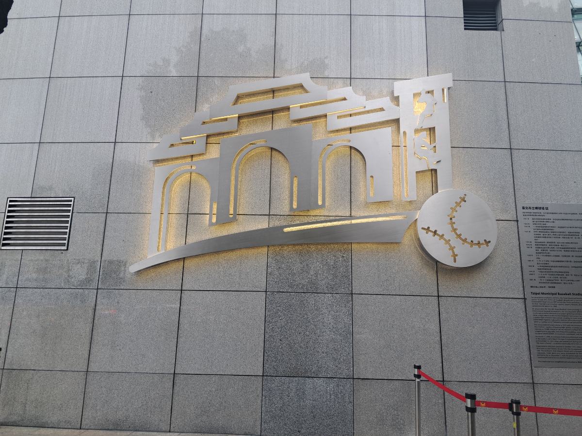 在台北小巨蛋牆上的紀念碑設計蘊含深厚情感：右側浮雕重現早年球迷「攀上燈塔看棒球」的經典畫面，為了一場球賽翻牆、爬高、踮腳張望的純粹熱血，正是市立棒球場最動人的風景。(影像由記者郭晉嘉拍攝)。
