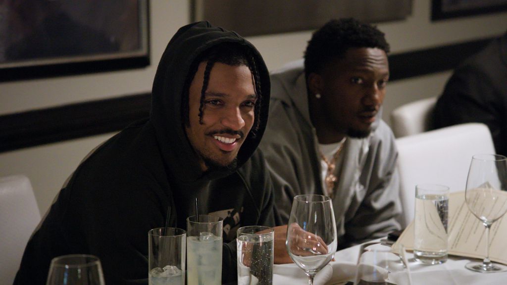"Two men sit at a restaurant table set with glasses, menus, and water. One man, wearing a hoodie, smiles while conversing. The setting is casual and relaxed with contemporary decor."