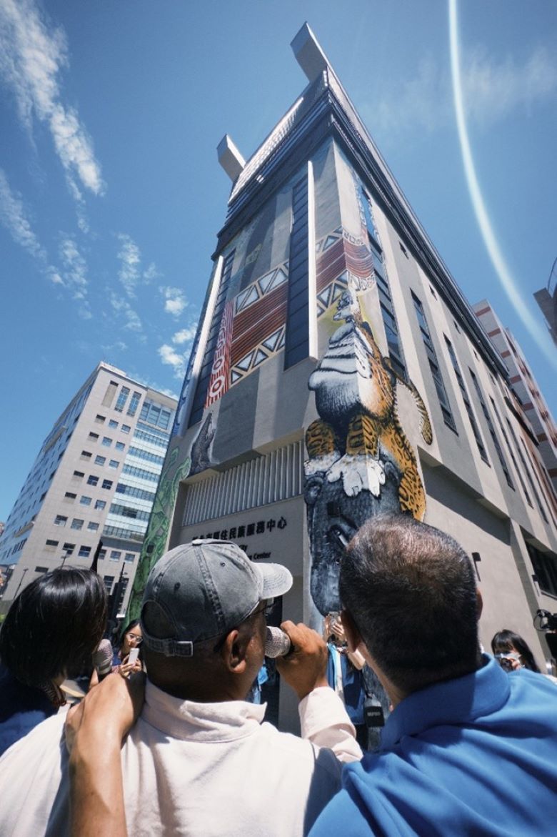 三重北原中心9月底開館 外牆Q版雲豹彩繪搶先亮相 48 《圖說》北原中心外牆彩繪藝術家杜寒菘(戴帽者)向侯友宜市長(圖右)解說創作理念。〈原民局提供〉