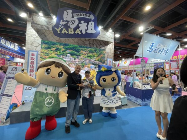 2026台北國際夏季旅展馬祖主題館舉辦互動問答活動，民眾熱情參與並成功答題，開心獲得精美馬祖特色贈品，在吉祥物陪伴下合影留念，現場氣氛熱絡，吸引不少民眾駐足參與。（朱立君攝）